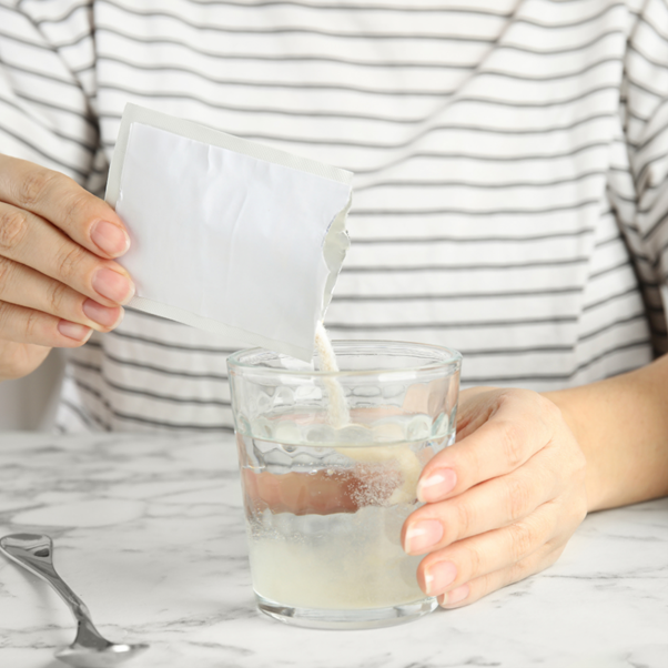 Close up image of a female wearing a white top with grey horizonal stripes emptying a sachet packet of vitamins into a small glass of water.