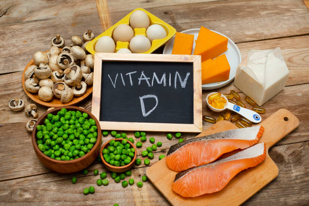 Various foods rich in Vitamin D, including eggs, cheese, mushrooms, salmon, green peas, butter, oil capsules, and milk, displayed on a wooden table with a chalkboard labeled "Vitamin D.