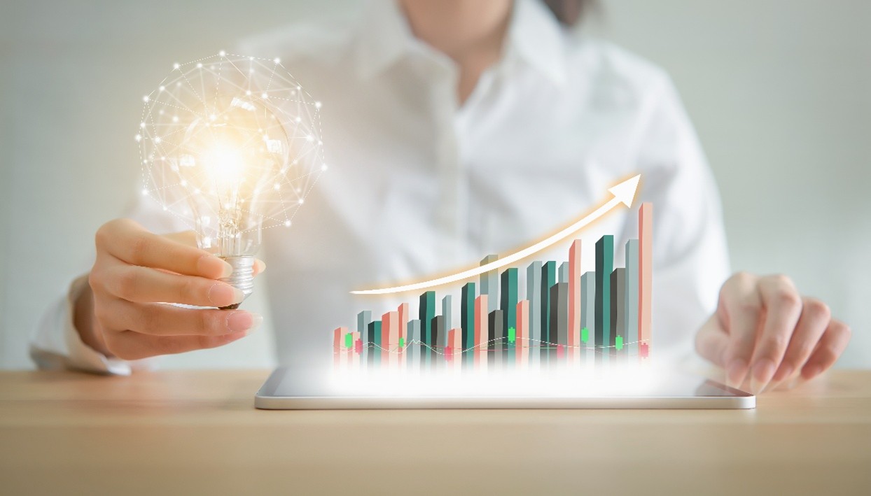 A person holds a glowing light bulb next to a rising bar graph projected from a tablet, symbolizing innovation and business growth.