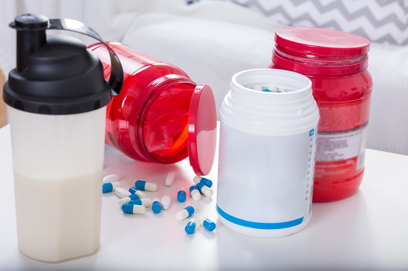A shaker bottle with a white liquid, two red supplement containers, and a white pill bottle with blue and white capsules scattered on a table.