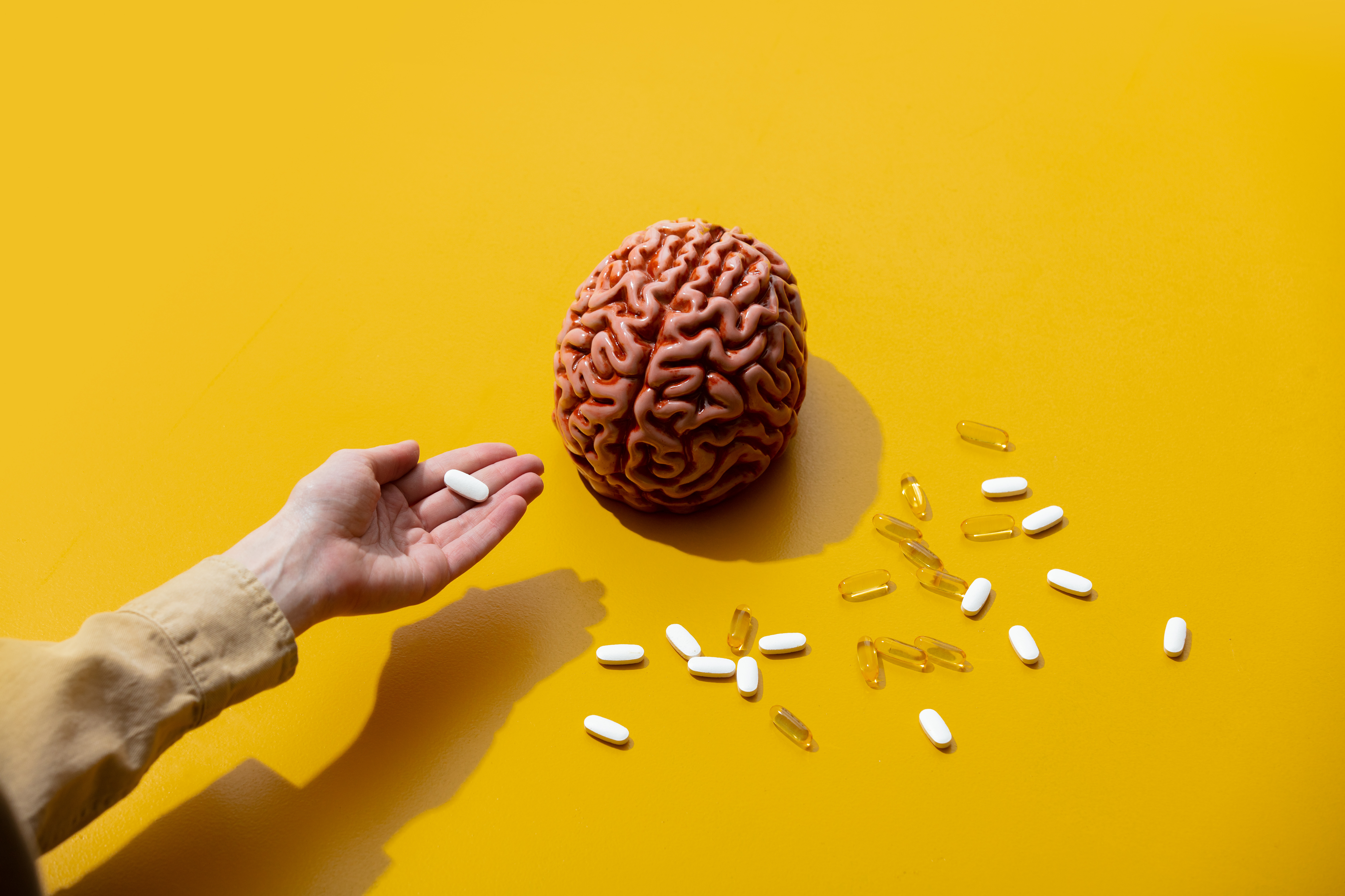 A hand holds a white pill near a model of a brain and scattered pills and capsules on a yellow background.