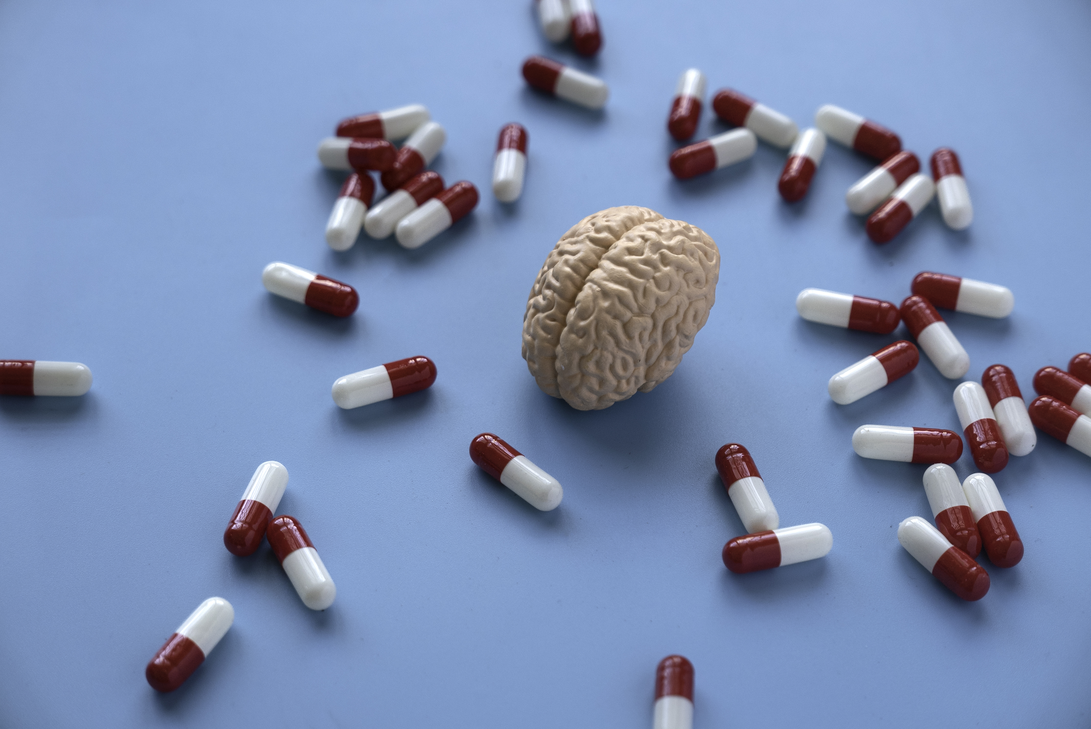 A small model brain sits on a blue surface surrounded by scattered red and white capsules.