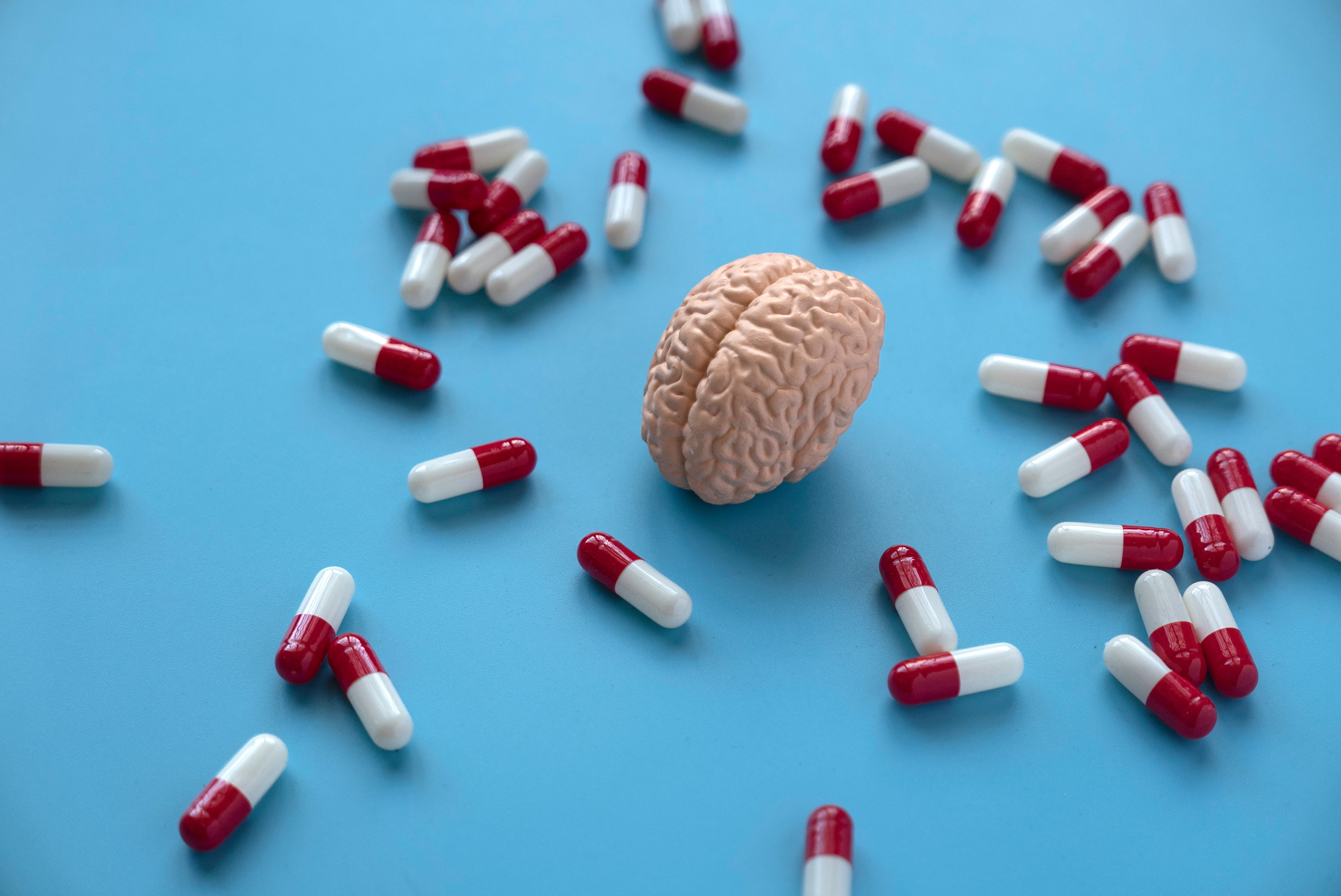 A small model brain sits on a blue surface surrounded by scattered red and white capsules.