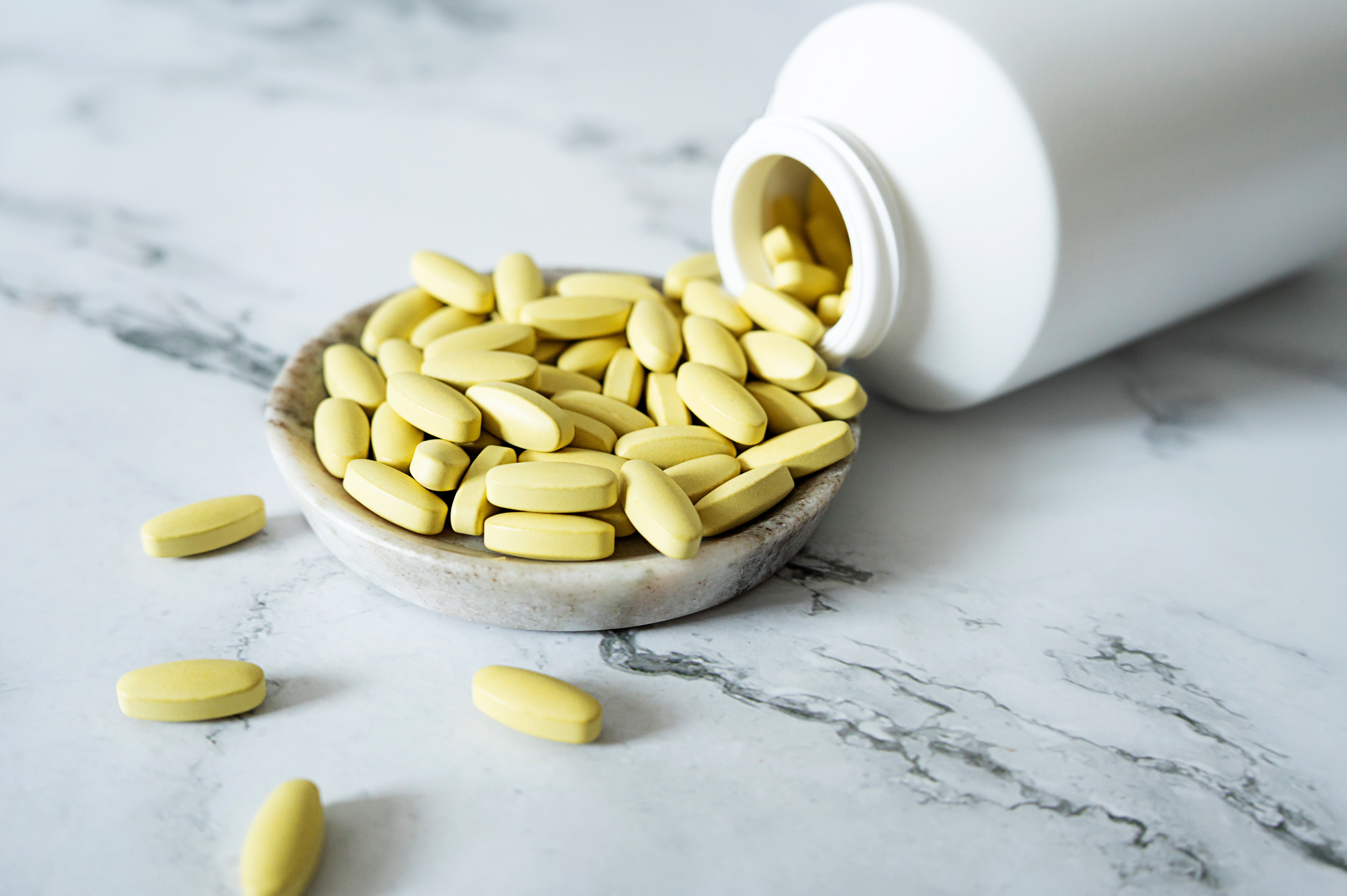Yellow oval pills spilling from a white plastic bottle onto a small marble dish.